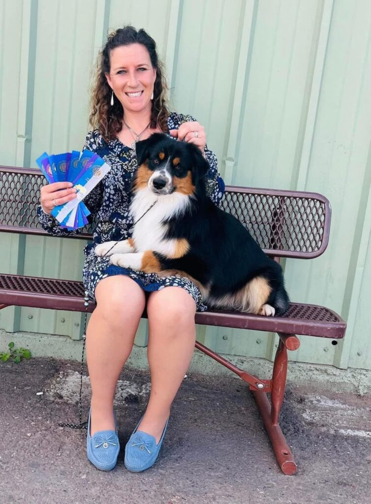 Laddie sitting on a bench beside his handler, who holds a fan of show ribbons